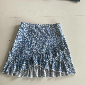 Blommig blå volangkjol från SHEIN - Supersöt blå kjol från SHEIN med vitt blommönster och volangdetalj längst ner. Kjolen är kort och har en omlott-look framtill. Perfekt för dig som gillar färgglada och lekfulla plagg med lite extra detaljer.