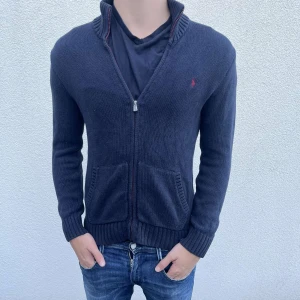 Mörkblå stickad kofta från Ralph Lauren - Snygg mörkblå stickad kofta från Ralph Lauren med dragkedja och två framfickor. Klassisk röd logga broderad på bröstet och ribbade muddar vid ärmslut och krage. Perfekt för dig som gillar stilren och chill look.