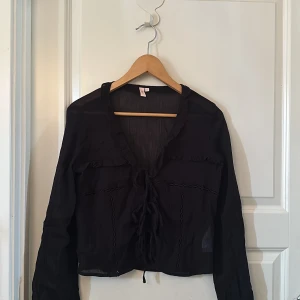 Svart blus med snörning från Nelly - Svart transparent blus från Nelly i storlek XS. Blusen har långa ärmar, v-ringning och snygga volangdetaljer längs halsen. Framtill finns snörning som ger en cool touch. Materialet är lätt och luftigt, perfekt för att styla med jeans eller kjol.