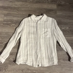 Vit randig linneskjorta från Zara - Vit skjorta från Zara i storlek M, tillverkad i linne. Skjortan har tunna vertikala ränder i grått, klassisk krage och knappstängning framtill. En bröstficka på vänster sida och långa ärmar med manschett. Perfekt för en chill och stilren look.