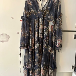 Blommig långklänning med volanger - Romantisk långklänning i mörkblått med blommigt mönster i beige, rosa och grönt. Klänningen har v-ringning, långa ärmar med knyt och volangdetaljer. Tunt, flowy material som ger en bohemisk känsla. Perfekt för dig som gillar en dreamy och chic stil.