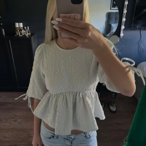 Vit blus med öppen rygg och knyt - Supersöt vit blus med strukturmönster, volang nertill och puffiga trekvartsärmar med knyt vid ärmslut. Baksidan är helt öppen med knytband upptill och i midjan. Perfekt för dig som gillar unika detaljer och romantisk stil.