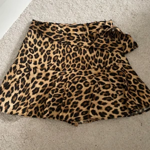 Leopardmönstrad kjol med volang från Zara - Snygg kortkjol med leopardmönster och volangdetalj upptill från Zara. Kjolen har omlottkänsla och knyts i midjan för en trendig look. Perfekt för dig som vill sticka ut och älskar djurmönster.