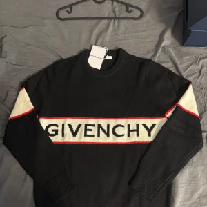 Svart stickad tröja från Givenchy - Svart stickad tröja från Givenchy med vit och röd rand över bröstet och armarna. Stor Givenchy-logga framtill. Klassisk rund halsringning och långa ärmar. Perfekt för dig som vill ha en stilren men ändå statement look.