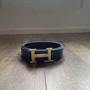 Mörkblått skärp med guldfärgat H-spänne - Snyggt mörkblå bälte med ikoniskt H-spänne i guldfärg. Bältet är gjort i slätt skinn och har vita kontrastsömmar längs kanterna. Perfekt accessoar för att lyfta vilken outfit som helst.