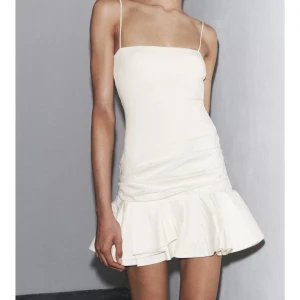 Vit kort klänning med volang Zara - Supersöt vit kort klänning med smala axelband och volang nertill. Klänningen har en rak, figurnära passform och ärmlös design. Perfekt för sommarens alla tillfällen när du vill känna dig extra stylish.