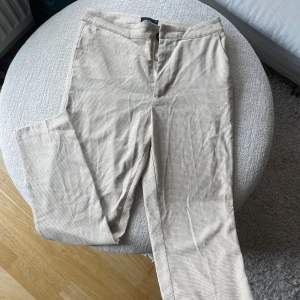 Beige rutiga kostymbyxor Gina Tricot - Snygga beige kostymbyxor från Gina Tricot i storlek 36. Byxorna har ett diskret rutigt mönster och rak passform med klassisk midja. Perfekta för en clean och stilren look. Stängs med dragkedja och knapp framtill.