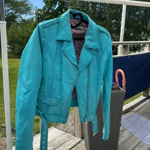 Turkos skinnjacka Jofama by Kenza - Snygg turkos skinnjacka från Jofama by Kenza med flätad detalj på axlarna, dragkedja framtill och bälte i midjan. Jackan har blommigt foder och coola metalldetaljer. Perfekt statement-piece för dig som vill sticka ut.