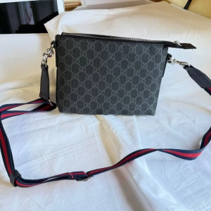 Svart axelväska från Gucci med GG-mönster - Snygg svart axelväska från Gucci med klassiskt GG-mönster över hela väskan. Väskan har en bred axelrem i marinblått och rött, silverfärgade metalldetaljer och dragkedja upptill. Tillverkad i canvas med detaljer i skinn och har ett innerfack med dragkedja.