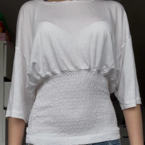 Vit topp från JUST Female med smock - Superfin vit topp från JUST Female med smockad midja som ger en snygg siluett. Modellen har trekvartsärmar och är gjord i mjuk bomull. Perfekt till jeans eller kjol för en clean och fräsch look. Finns en liten svag fläck som ej går bort men syns knappast🥰och köptes för 550kr. Pris kan diskuteras! Går för både XS, S 