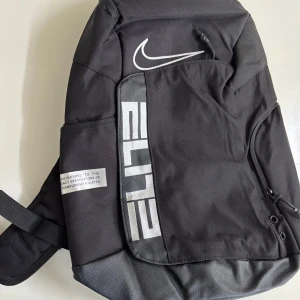 Nike Elite svart ryggsäck - Säljer en svart Nike Elite ryggsäck med stora vita loggor och coola detaljer på axelremmarna. Ryggsäcken har flera fack, dragkedjor och vadderade remmar för extra komfort. Perfekt för träning eller skola, med sportig och stilren look.