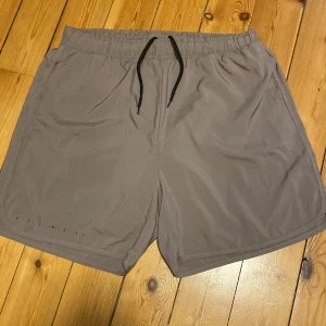 Grå träningsshorts med snörning - Snygga grå träningsshorts med elastisk midja och svart snörning. Shortsen har en loose passform och är tillverkade i ett lätt syntetmaterial som andas. Perfekta för gymmet eller löprundan. Diskret logga på baksidan upptill.