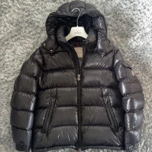 Svart pufferjacka från Moncler - Säljer en svart, glansig pufferjacka från Moncler med huva och dragkedja. Jackan har två snedställda fickor med dragkedja och en Moncler-logga på ärmen. Perfekt för kalla dagar och riktigt snygg till streetwear-stilen.