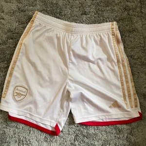 Arsenal fotbollsshorts - Snygga vita Arsenal shorts från Adidas med guldiga ränder längs sidorna och röd kant längst ner. Klassisk sportig look med elastisk midja och broderad Arsenal-logga på ena benet. Perfekta för fotboll eller träning.