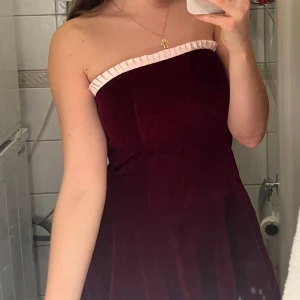 Vinröd sammetsklänning med snörning - Superfin vinröd sammetsklänning med axelbandslös design och söt ljusrosa volangkant upptill. Klänningen har snörning i ryggen och en utsvängd kjol som ger en riktigt drömmig siluett. Perfekt för jul eller andra speciella tillfällen!
