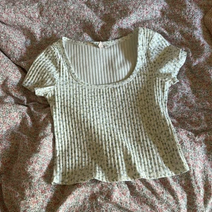 Blommig ribbad topp från Nelly - Supersöt croppad topp från Nelly i vitt med små blå  blommor med en grön stam. Toppen är ribbad, har korta ärmar och en rundad, lite djupare urringning. Perfekt för en fräsch och somrig look!