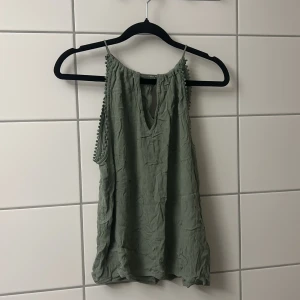 Grön ärmlös linnetopp med v-ringning - Snygg ljusgrön linnetopp med v-ringning och dekorativa små bollar längs axelbanden. Toppen är ärmlös och har en avslappnad, loose passform. Perfekt för varma dagar och enkel att styla med jeans eller shorts.