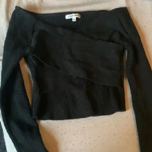 Svart ribbad offshoulder topp Cherry Koko - Säljer en svart ribbad offshoulder topp från Cherry Koko. Toppen har lång ärm, croppad passform och en snygg omlott-detalj framtill. Perfekt för dig som gillar stilrena och trendiga plagg med lite edge. Tryck ej köp nu