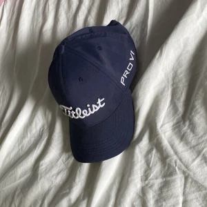 Mörkblå Titleist keps Tour Performance - Snygg mörkblå keps från Titleist med broderad logga framtill och på sidan. Justerbar rem baktill med rödvit Titleist-detalj. Klassisk böjd skärm och ventilerande hål. Perfekt för golf eller sportiga dagar.