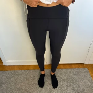 Svarta leggings från Lululemon - Snygga svarta leggings från Lululemon med hög midja och praktiska sidfickor. De är tighta och sitter perfekt för träning eller chill. Tillverkade i ett stretchigt syntetmaterial som känns mjukt mot huden. Klassisk Lululemon-logga bak på linningen.