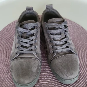 Grå mockasneakers från Christian Louboutin - Snygga grå sneakers från Christian Louboutin i mjuk mocka med klassisk röd sula. Skorna har rund tå, platt sula och grå snörning. Perfekta för dig som vill ha en stilren och exklusiv look med en lyxig touch.