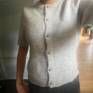 Ljusgrå stickad kofta med pärlknappar - Superfin ljusgrå stickad kofta med korta ärmar och snygga pärlknappar framtill. Modellen är lite croppad och har ribbade muddar vid ärmslut och nederkant. Perfekt att slänga över en topp för en chill men ändå stilsäker look.