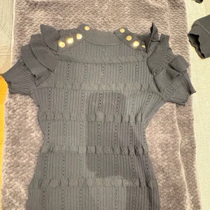 Svart ribbad kortklänning May byshiningstar - Supersnygg svart ribbad kortklänning från May byshiningstar. Klänningen har coola volangdetaljer på axlarna, korta ärmar och guldknappar vid axlarna. Stickad struktur med hålmönster ger en trendig look. Perfekt för dig som gillar unika detaljer! 