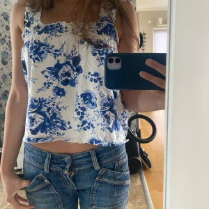 Blommig blåvit linnetopp - Supersöt linnetopp med blåa blommor på vit botten. Toppen har tjocka axelband, rynkade detaljer upptill och en luftig passform. Perfekt för varma dagar och ger en fräsch sommarkänsla.