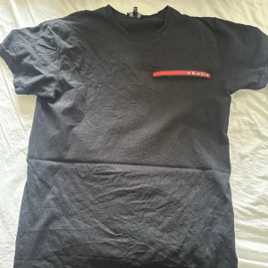 Svart t-shirt från Prada - Svart t-shirt från Prada med klassisk passform och rund hals. På bröstet finns en röd detalj med Prada-loggan.