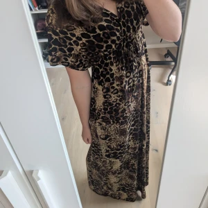 Långklänning med leopardmönster - Säljer en maxiklänning med v-ringning och snygg leopardmönstrad design. Klänningen har korta ärmar, knytning under bysten och är i mjukt, följsamt material. Perfekt för dig som vill sticka ut med ett djurmönster och en skön passform.