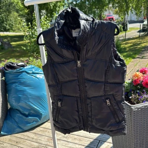 Svart dunväst från Rosetti Riding - Snygg svart dunväst från Rosetti Riding med hög krage och två dragkedjefickor framtill. Västen har quiltad design och stängs med en rejäl dragkedja. Perfekt för dig som gillar en sportig och clean look.