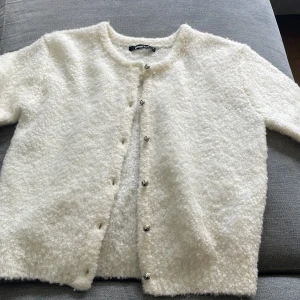 Vit kortärmad kofta från Gina Tricot - Supermjuk och fluffig vit kofta från Gina Tricot med korta ärmar och silvriga knappar framtill. Koftan har en rund halsringning och är stickad i ett mysigt material som känns lyxigt och trendigt. Perfekt att slänga över en topp för en chill look.