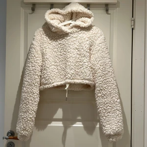 Beige/vit teddy hoodie med huva - Supermjuk och fluffig beige/vit teddy hoodie med croppad passform och snörning i midjan. Tröjan har långärmade ärmar och stor huva, perfekt för kalla dagar när du vill vara extra cozy.