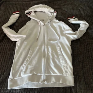  Moncler kofta  - Snygg vit kofta  från Moncler med hel dragkedja, huva och fickor framtill. Ärmar och huva har stilrena ränder i rött, blått och vitt. Moncler-logga på ena ärmen. Tillverkad i mjuk bomull för en skön känsla.