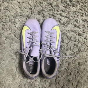 Ljuslila Nike fotbollsskor med neon - Snygga fotbollsskor från Nike i ljuslila med neonfärgade detaljer och klassisk Swoosh. Skorna har snörning, platt sula med dobbar och är tillverkade i syntetmaterial. Perfekta för dig som vill sticka ut på planen med stilrena och moderna skor.