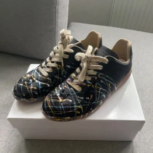 Maison margela sneakers med färgstänk - Unika svarta sneakers från Valentino Garavani med coolt färgstänkt mönster i blått, gult och vitt. Skorna har beige snörning och bruna gummisulor med grepp. Perfekta för dig som vill sticka ut med din stil.