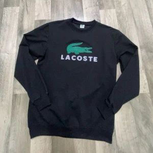 Svart Lacoste sweatshirt med logga - Svart sweatshirt från Lacoste med stor grön krokodil och vit Lacoste-text på bröstet. Tröjan har rund hals, långa ärmar och ribbade muddar. Perfekt för dig som gillar klassisk streetstyle och vill ha en tröja med tydlig logga.