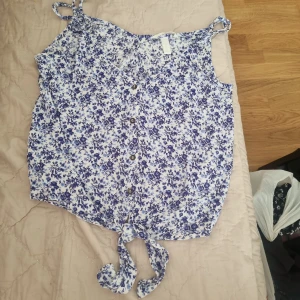 Blommig knyt-topp i blått och vitt - Supersöt blommig linnetopp i vitt med blått mönster. Toppen har knyt framtill, knappar längs hela framsidan och smala axelband. Perfekt för varma dagar och ger en fräsch vibe till din outfit. 