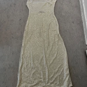 Glittrig långklänning i champagne - Supersnygg långklänning i champagnefärg med glittrigt tyg som verkligen fångar ljuset. Klänningen har en figurnära passform och är ärmlös, perfekt för att sticka ut på festen. Materialet känns lyxigt och skimrande.