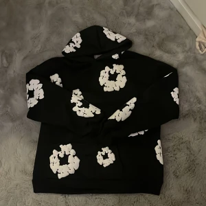 Denim Tears svart hoodie med vita blommor - Svart hoodie från Denim Tears med stora vita blommönster över hela tröjan och huva. Tröjan har en klassisk känguruficka framtill och är tillverkad i mjuk bomull. Perfekt statement-plagg för dig som vill sticka ut. Använd 1-2 gånger så det är bra skick💞