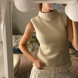 Beige ärmlös topp med fransdetalj - Superfin beige ärmlös topp med hög krage och fluffig textur. Toppen har en snygg fransdetalj längst ner som ger en cool touch. Perfekt att styla med jeans eller kjol för en trendig look.
