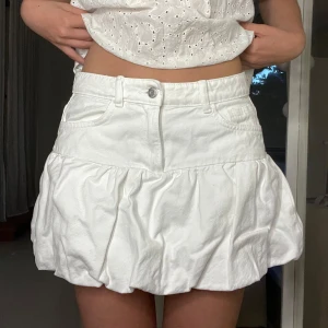 Vit volangkjol i jeansstil - Supersöt vit kortkjol med volang och jeansdetaljer. Kjolen har fickor fram, bälteshällor och dragkedja med knapp. Perfekt för sommarens alla tillfällen och ger en fräsch vibe till din outfit.