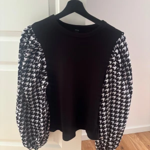 Svart tröja med mönstrade ärmar från SHEIN - Svart tröja från SHEIN med ballongärmar i svartvitt hundtandsmönster. Ärmarna är långa och ger en trendig look. Perfekt för dig som vill sticka ut lite extra med coola kontraster. Materialet känns mjukt och bekvämt mot huden.