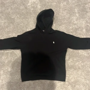 Svart hoodie från Polo Ralph Lauren - Svart hoodie från Polo Ralph Lauren med klassisk vit broderad logga på bröstet. Tröjan har huva, lång ärm och känguruficka framtill. Tillverkad i mjuk bomull, perfekt för en chill och stilren look.