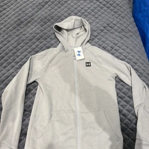 Grå hoodiejacka från Under Armour - Snygg grå hoodiejacka från Under Armour med dragkedja hela vägen, huva och två stora fickor framtill. Jackan har en stilren look med diskret logga på bröstet och är tillverkad i mjukt bomullsmaterial. Perfekt för chill dagar eller till träningen.