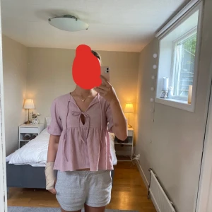 Ljusrosa blus med knyt och puffärm - Supersöt ljusrosa blus med puffiga korta ärmar och två knytband framtill. Blusen har en lös passform och är tillverkad i ett mjukt, strukturerat bomullstyg. Perfekt för en chill och trendig look. Storlek S och från Lager 157.