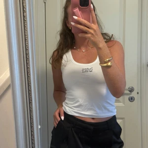 Vit linnetopp med broderad text - Säljer en vit linnetopp i bomull med broderad svart text på bröstet: 'The obvious is obviously not that obvious'. Modellen är ärmlös och har en klassisk rund halsringning. Perfekt till sommaren och enkel att styla med jeans eller kjol.