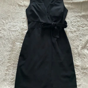 Svart  klänning med knyt - Stilren svart kortklänning med omlottkänsla och knytband i midjan. Ärmlös modell med v-ringad hals och dragkedja i ryggen. Perfekt för dig som gillar en klassisk look med modern twist.