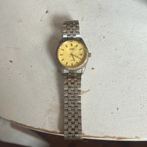 Visa Quartz klocka med gul urtavla - Stilren klocka från Visa med klassisk länk i silverfärgad metall och en gul urtavla. Klockan har datumvisning och tydliga indexmarkeringar. Japansk kvartsrörelse och räfflad kant runt urtavlan ger en cool retrovibe.