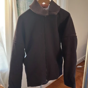 Svart jacka Bruno Pieters x Weekday - Säljer en svart jacka från Bruno Pieters x Weekday med hög ribbad krage och hel dragkedja framtill. Jackan har en minimalistisk design, raka linjer och är tillverkad i ett mjukt material som känns både stilrent och bekvämt. Perfekt för dig som gillar clean och modern look.